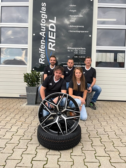 Das Team von Reifen-Autoglas Riedl vor der Werkstatt mit einem Komplettrad  spezialisiert auf Reifen, Felgen, Autoglas und Steinschlagreparatur." 